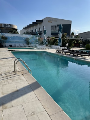 A clear blue swimming pool surrounded by a tiled deck and several sun loungers with parasols. The modern hotel building in the background features balconies and a small garden area beside the pool. The sky is clear, suggesting a sunny day, and the overall setting appears serene and inviting.