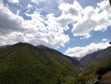 Lush green hills with dense forests fill the foreground, stretching towards the horizon, where snow-capped mountains rise majestically. The sky above is partly cloudy with patches of blue, creating a serene and picturesque landscape.