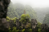 A serene landscape featuring lush green trees and rocky cliffs with a small pagoda standing prominently among the foliage. The scene is enveloped in a gentle mist, enhancing the tranquility and mystery of the natural surroundings.