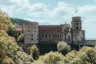 A historic, partially ruined castle with prominent stone towers and walls is surrounded by lush greenery. The structure features medieval architecture, including tall, narrow windows. A small flag is visible atop one of the towers, fluttering gently in a light breeze. The background includes a lightly clouded sky and distant hills.