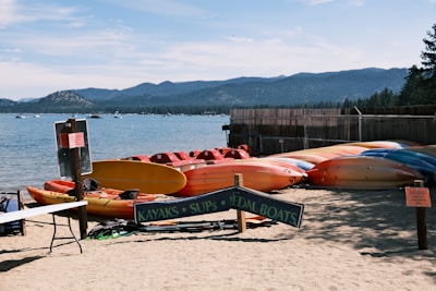 A sandy beach with multiple brightly colored kayaks and pedal boats are displayed in neat rows under a sign. The lake is calm and expansive, with gentle waves lapping at the shore. In the distance, forested mountains rise against a clear blue sky. Signs and small tables are scattered around, indicating a rental or recreation area.