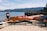 A sandy beach with multiple brightly colored kayaks and pedal boats are displayed in neat rows under a sign. The lake is calm and expansive, with gentle waves lapping at the shore. In the distance, forested mountains rise against a clear blue sky. Signs and small tables are scattered around, indicating a rental or recreation area.