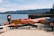 A sandy beach with multiple brightly colored kayaks and pedal boats are displayed in neat rows under a sign. The lake is calm and expansive, with gentle waves lapping at the shore. In the distance, forested mountains rise against a clear blue sky. Signs and small tables are scattered around, indicating a rental or recreation area.