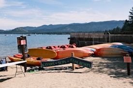 A sandy beach with multiple brightly colored kayaks and pedal boats are displayed in neat rows under a sign. The lake is calm and expansive, with gentle waves lapping at the shore. In the distance, forested mountains rise against a clear blue sky. Signs and small tables are scattered around, indicating a rental or recreation area.