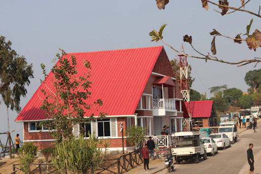 A modern house with a distinctive red triangular roof and white accents. The building features several windows and a balcony, surrounded by a landscaped area with trees and bushes. There are people walking and vehicles, including cars and trucks, parked along the adjacent road. The scene is set in a rural or suburban area with a clear sky.