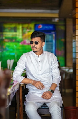 A man wearing sunglasses and a traditional white kurta sits confidently in a restaurant setting with a colorful, blurred background. The ambiance seems casual and relaxed.