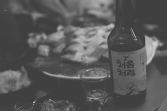 A close-up of food on a table with an opened bottle featuring Korean text prominently displayed in the foreground. The background shows plates with various dishes, suggesting a dining setting.