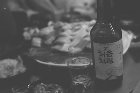A close-up of food on a table with an opened bottle featuring Korean text prominently displayed in the foreground. The background shows plates with various dishes, suggesting a dining setting.