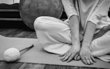 A person sits cross-legged on a mat, with hands resting on the floor. The person is dressed in light, loose clothing. Nearby, there is a large round drum and a drumstick with a soft mallet head.