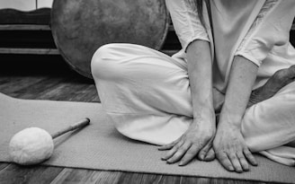 A person sits cross-legged on a mat, with hands resting on the floor. The person is dressed in light, loose clothing. Nearby, there is a large round drum and a drumstick with a soft mallet head.