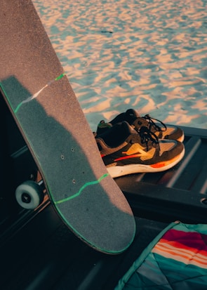 A skateboard with a gray grip tape and green accents rests against a vehicle's interior. Nearby, a pair of black and white sneakers with orange and red details are placed. The background shows a sandy texture, suggesting a beach setting. A multicolored fabric with stripes is visible on the lower right side.