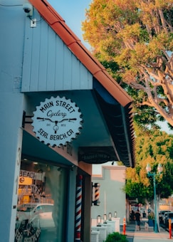 A quaint street scene featuring the front of a building with a sign for a cycling shop in Seal Beach, California. The building has a blue and white exterior with a distinctive orange roof edge. Below the cycling shop sign, there is a traditional barber pole, indicating a nearby barbershop. The street is lined with trees, one of which is prominently visible, providing greenery against the backdrop of early evening sky.
