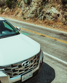 A white Hyundai car is parked near a rocky hillside on a two-lane road. The license plate is visible on the front of the vehicle, and the surrounding landscape includes rough terrain with some vegetation.