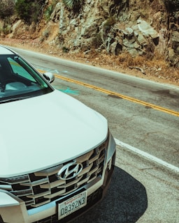 A white Hyundai car is parked near a rocky hillside on a two-lane road. The license plate is visible on the front of the vehicle, and the surrounding landscape includes rough terrain with some vegetation.