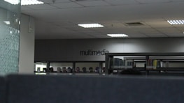 The image depicts an indoor space with bookshelves lined against a wall. The word 'multimedia' is visible on the far wall, indicating that this section could be related to multimedia resources. The ceiling has recessed lighting, and the overall tone of the space is modern and minimalistic.
