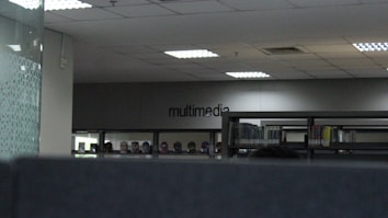 The image depicts an indoor space with bookshelves lined against a wall. The word 'multimedia' is visible on the far wall, indicating that this section could be related to multimedia resources. The ceiling has recessed lighting, and the overall tone of the space is modern and minimalistic.