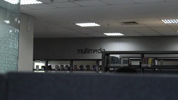 The image depicts an indoor space with bookshelves lined against a wall. The word 'multimedia' is visible on the far wall, indicating that this section could be related to multimedia resources. The ceiling has recessed lighting, and the overall tone of the space is modern and minimalistic.