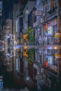 A nighttime urban scene features a street lined with buildings adorned with neon signs and lights. The reflection of the entire scene is visible in a puddle on the ground, doubling the visual effect. A person holding a transparent umbrella walks along the street, creating a sense of calm amidst the vibrant city life.