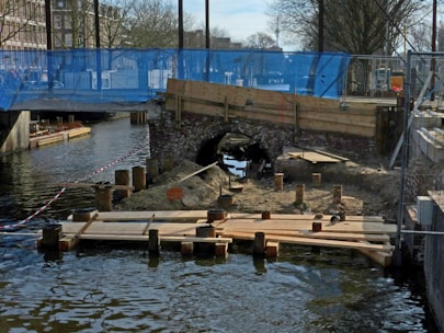 A construction site near a canal features wooden planks placed on top of posts in the water, with a small archway constructed of bricks in the background. The area is fenced off with blue mesh and red-striped tape. Buildings and leafless trees are visible in the distance, suggesting an urban setting.