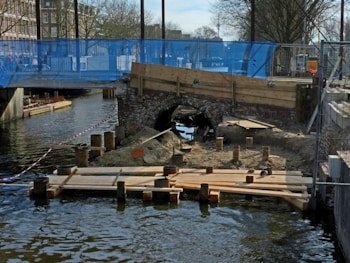 A construction site near a canal features wooden planks placed on top of posts in the water, with a small archway constructed of bricks in the background. The area is fenced off with blue mesh and red-striped tape. Buildings and leafless trees are visible in the distance, suggesting an urban setting.