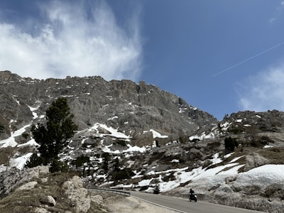 A winding road cuts through a mountainous landscape with patches of snow scattered across rocky terrain. A lone motorbike travels along the road under a bright blue sky, accompanied by some clouds. The rugged mountains tower in the background, with sparse vegetation and a few evergreen trees.