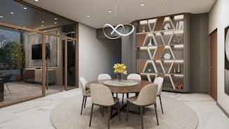 A modern dining area with a round wooden table and six upholstered chairs on a textured circular rug. Above the table hangs a unique, contemporary light fixture. The room features a large, geometric shelving unit with decorative items and books. Large glass doors lead to another room with visible indoor plants and a television.