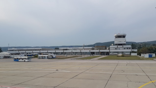 An airport runway with a terminal building and a control tower in the background. Buses and equipment are visible on the tarmac, and the surrounding landscape includes trees and distant hills under a cloudy sky.
