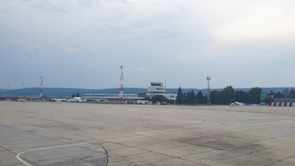 A large, open tarmac area is situated in the foreground with visible markings and paving lines. In the distance, there is an airport terminal building including a control tower. Several red and white communication towers are scattered throughout the scene. Some small service vehicles are parked near the buildings. The horizon reveals a range of hills under a cloudy sky.