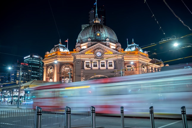 A grand historic train station is illuminated with warm lights against a night sky. A tram speeds by in the foreground, creating a motion blur effect. The station features a prominent clock and domed roof, surrounded by modern city buildings.