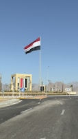 A large flag with red, white, and black stripes is flying atop a tall flagpole against a clear blue sky. The flag is surrounded by a roundabout with a landscaped area and a decorative structure featuring the same colors as the flag. The background features distant mountains, and the road has visible traffic signs and direction arrows.
