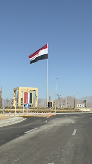 A large flag with red, white, and black stripes is flying atop a tall flagpole against a clear blue sky. The flag is surrounded by a roundabout with a landscaped area and a decorative structure featuring the same colors as the flag. The background features distant mountains, and the road has visible traffic signs and direction arrows.