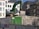 A large green excavator is positioned on a mound of rubble in an urban construction site. Surrounding the machinery are tall residential buildings with multiple windows and a visible rooftop. The scene includes a metal fence and a green control box with labels.