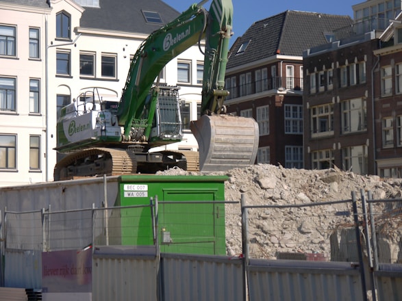 A large green excavator is positioned on a mound of rubble in an urban construction site. Surrounding the machinery are tall residential buildings with multiple windows and a visible rooftop. The scene includes a metal fence and a green control box with labels.