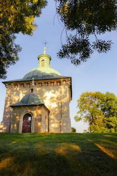 A small chapel with a dome roof situated on a grassy hill. Sunlight creates dappled patterns on the building's stone exterior. Lush green trees partially frame the scene under a clear blue sky. A cross sits at the top of the dome.