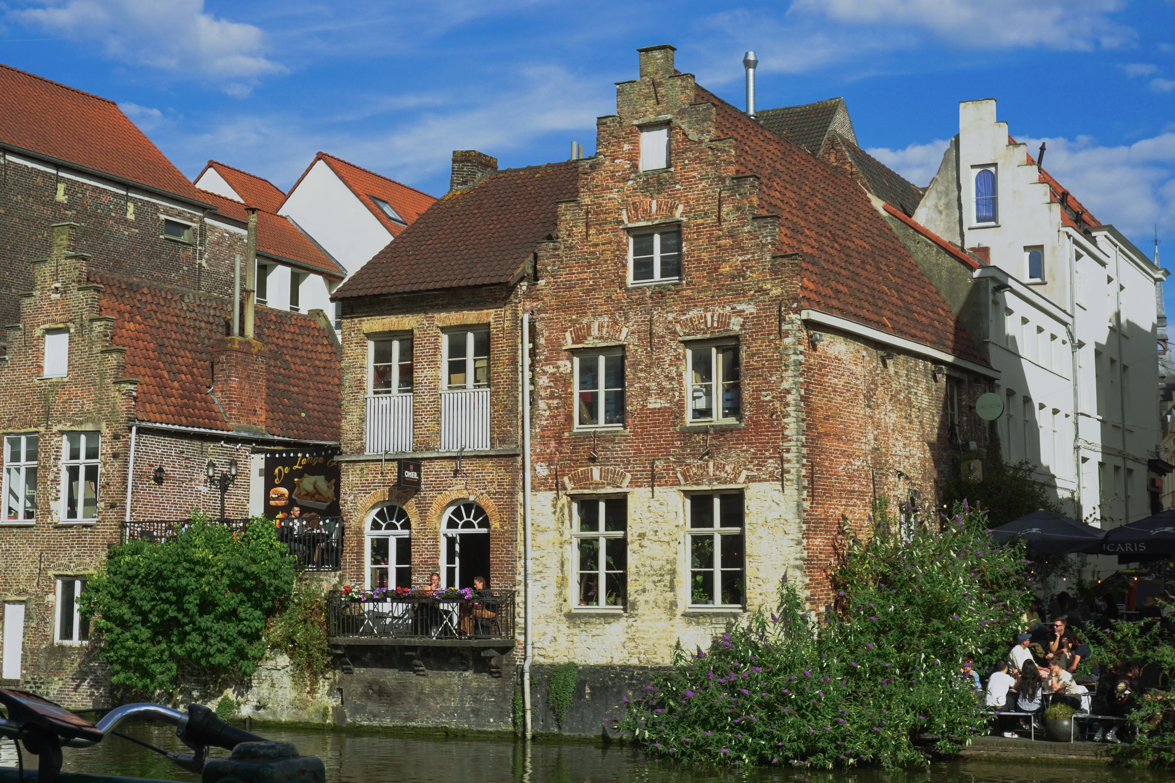 A weathered brick stepped-gable house on a canal, with an outdoor cafe terrace and greenery in sunlight.