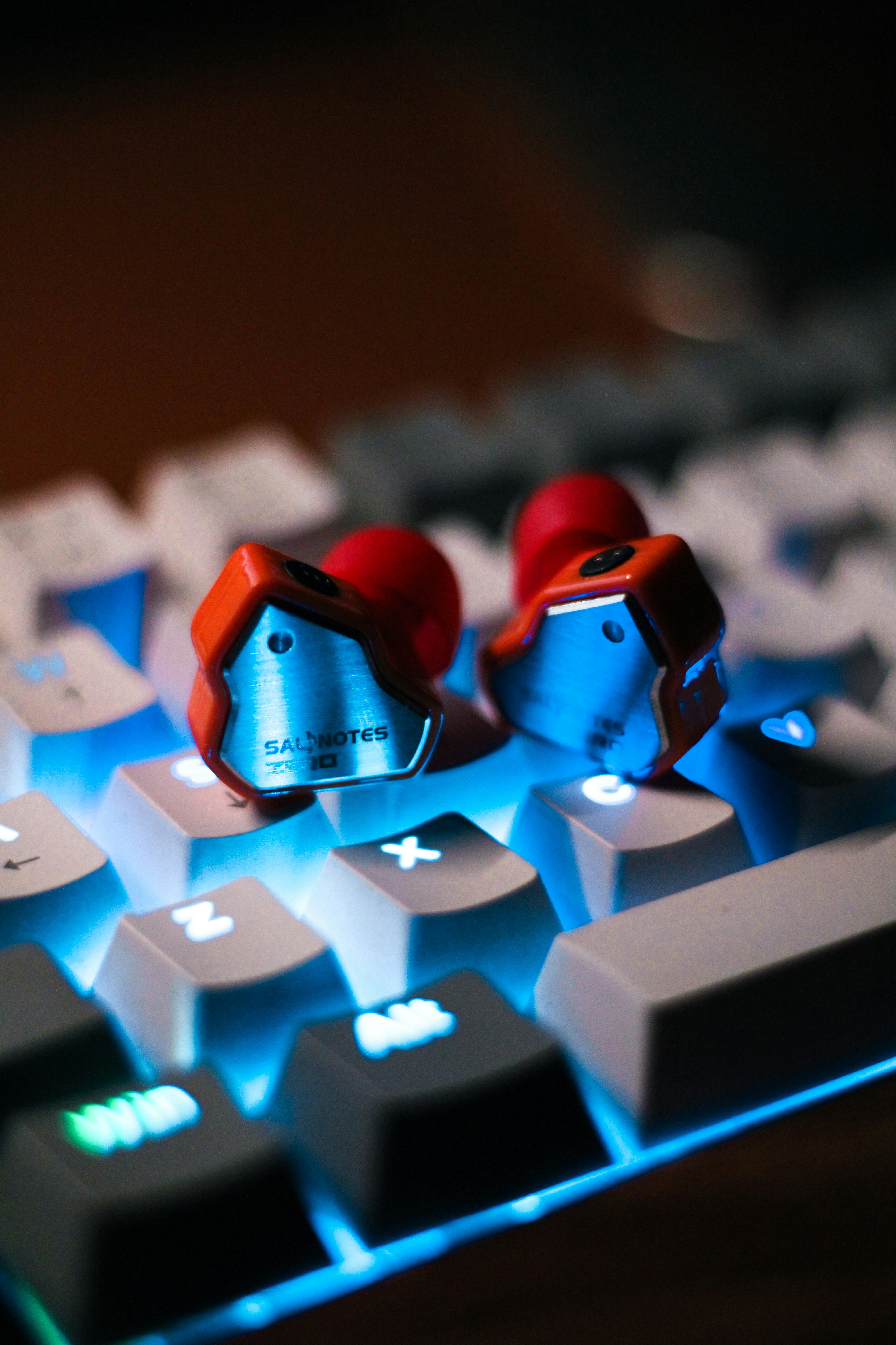 A pair of earphones rests on a backlit keyboard. The earphones have a metallic exterior and prominent red accents. The keyboard keys are illuminated with a bright blue light, creating a contrasting background.