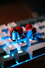 A pair of earphones rests on a backlit keyboard. The earphones have a metallic exterior and prominent red accents. The keyboard keys are illuminated with a bright blue light, creating a contrasting background.