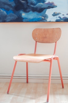 A wooden chair with a natural finish seat and backrest is supported by metal legs painted in a soft coral color. It stands on a light wood floor against a neutral wall. Above the chair, part of a painting featuring dark and light blue clouds is visible.