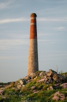 A tall, cylindrical stone chimney rises against a clear sky, standing on a grassy hill dotted with rocks and pink wildflowers. The structure is made of bricks in the upper section and stone in the lower section.