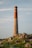 A tall, cylindrical stone chimney rises against a clear sky, standing on a grassy hill dotted with rocks and pink wildflowers. The structure is made of bricks in the upper section and stone in the lower section.