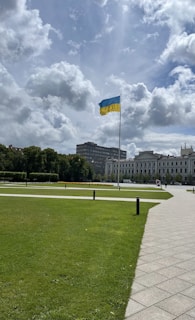 A large national flag is raised prominently in a spacious park area. The grass is neatly trimmed, and a clear pathway is visible. In the background, a stately building, likely governmental or historical, stands against a partly cloudy sky.