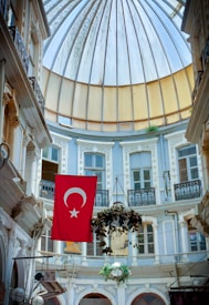 An ornate indoor courtyard with a high, arched glass ceiling. The central focus is a Turkish flag hanging prominently, surrounded by elegant, historical architecture featuring balconies and decorative moldings. A hanging plant adds greenery to the otherwise pastel and neutral surroundings.
