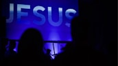 A large, illuminated word 'JESUS' is displayed prominently against a blue background. Silhouettes of people are seen in the foreground, suggesting a gathering or event.
