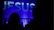 A large, illuminated word 'JESUS' is displayed prominently against a blue background. Silhouettes of people are seen in the foreground, suggesting a gathering or event.