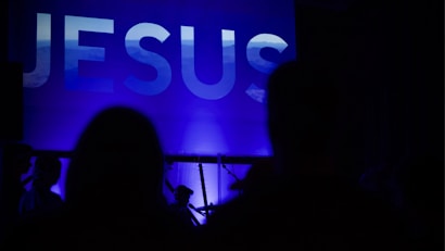 A large, illuminated word 'JESUS' is displayed prominently against a blue background. Silhouettes of people are seen in the foreground, suggesting a gathering or event.