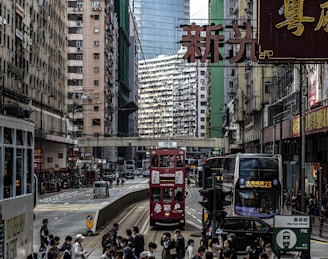 A bustling urban street with a red double-decker tram and a modern double-decker bus. High-rise residential and commercial buildings line both sides of the street. Numerous people can be seen commuting or waiting, contributing to a lively city atmosphere. Several neon signs with Chinese characters and various advertisements are prominently displayed.