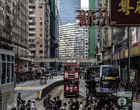 A bustling urban street with a red double-decker tram and a modern double-decker bus. High-rise residential and commercial buildings line both sides of the street. Numerous people can be seen commuting or waiting, contributing to a lively city atmosphere. Several neon signs with Chinese characters and various advertisements are prominently displayed.