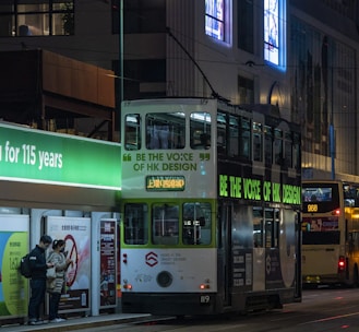 A night scene of a city street featuring a double-decker tram with advertisements promoting the 'Be the Voice of HK Design' campaign. The tram is stopped at a tram station with a glowing green sign. A couple of people stand at the station looking at their phones. In the background, another bus is visible, along with illuminated billboards and a building facade.