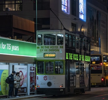 A night scene of a city street featuring a double-decker tram with advertisements promoting the 'Be the Voice of HK Design' campaign. The tram is stopped at a tram station with a glowing green sign. A couple of people stand at the station looking at their phones. In the background, another bus is visible, along with illuminated billboards and a building facade.