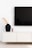 A minimalist interior scene features a large, black flat-screen television mounted on a white wall above a sleek, white modern console table. On the left side of the console sits a tall, black ceramic vase with long, wavy dried grasses, next to a small, cylindrical candle holder.